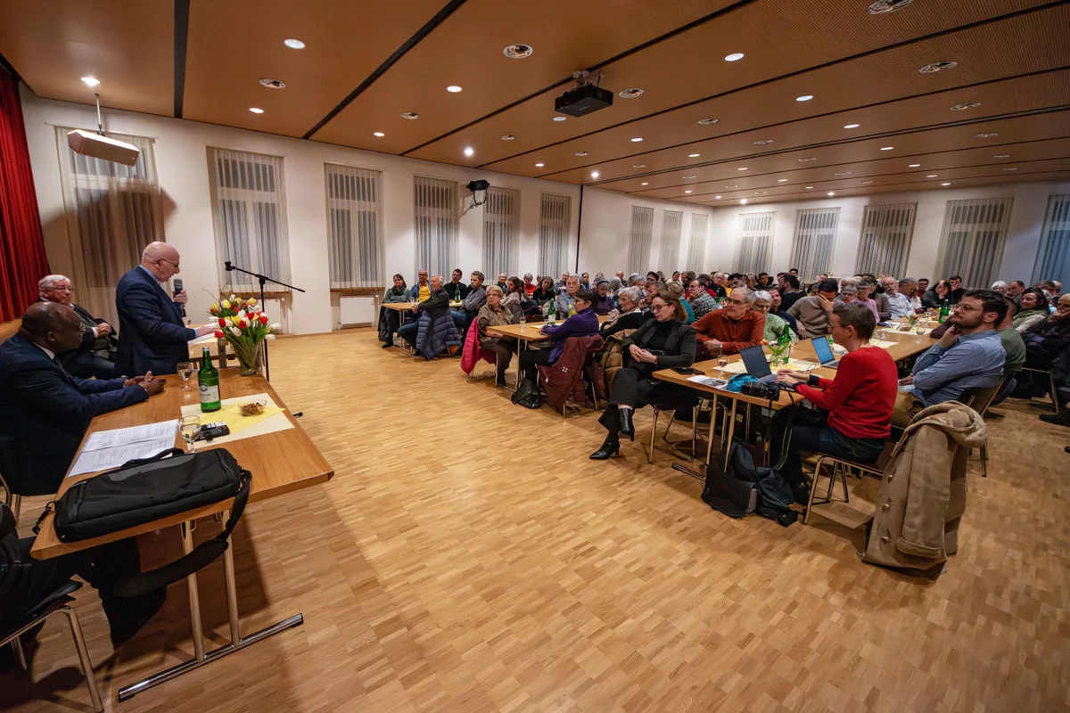 Man sieht einen Raum im Weitwinkel mit vielen Gästen, die längs an Tischen sitzen. 